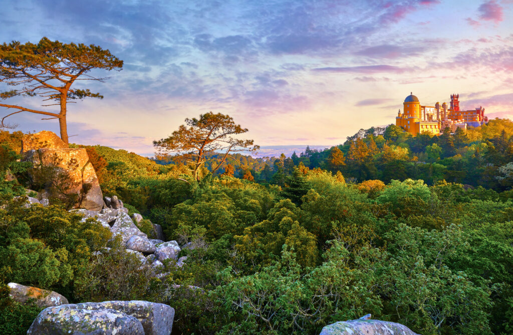 Reise Programm Sintra 2027 4 Kulturreisen Sintra 2027: Palácio Nacional da Pena auf bewaldetem Hügel, farbenprächtig im Abendlicht mit Granitfelsen im Vordergrund. Gartenreisen 2027 Termine sind verfügbar.