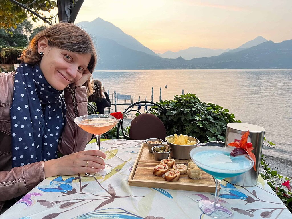 Reise Programm Comersee 2026 4 Aperitivo-Tisch mit Cocktails und Antipasti auf einer Uferterrasse am Comersee bei Sonnenuntergang, Bergpanorama im Hintergrund – Kulturzelt Reisen Kultur- und Gartenreise Comersee 2026