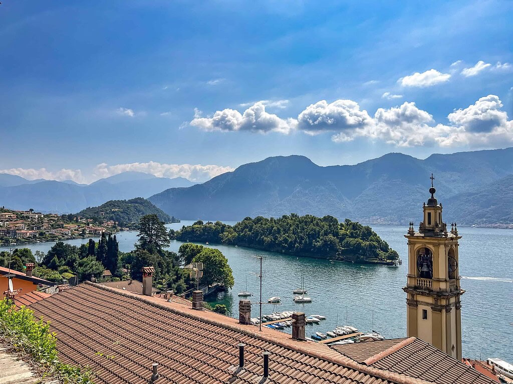 Comersee über die Reise 2026 9 Blick über die Dächer auf die Isola Comacina im Comersee, eingerahmt von einer barocken Kampanile und den Bergen der Lombardei – Kulturzelt Reisen Kultur- und Gartenreise Comersee 2027
