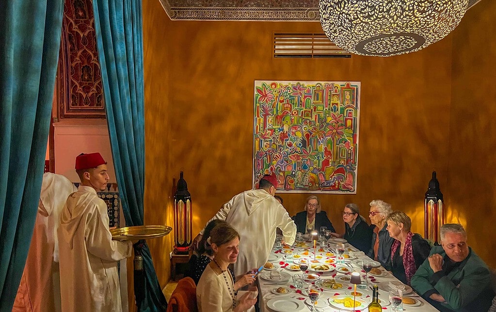 KULTURZELT REISEN 15 Reisegruppe an einer festlich gedeckten Tafel in einem Riad-Salon mit ockerfarbenen Wänden, Kellner in weissen Gewändern und Fes servieren
